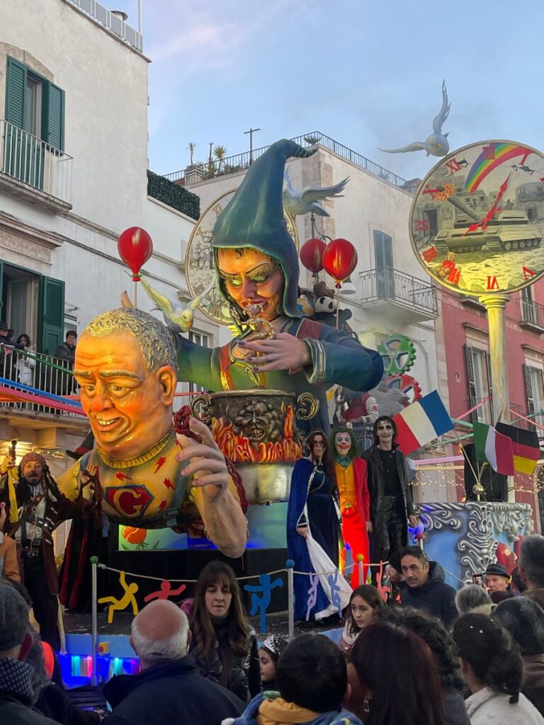 Carro colorato del Carnevale di Putignano con figure di clown giganti e palloncini rossi. Intorno ci sono persone in costume. Le bandiere e un orologio aggiungono festività. Scena gioiosa e vivace.