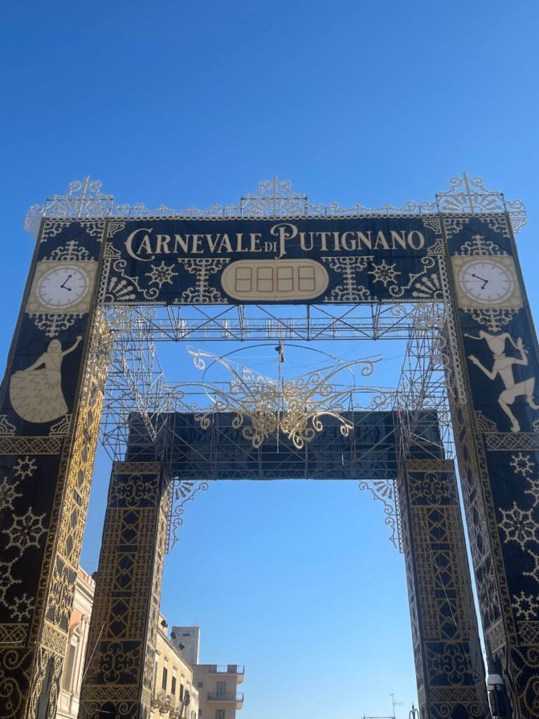 Arco ornato con disegni intricati e testo "Carnevale di Putignano" contro un cielo azzurro. Due orologi su entrambi i lati migliorano l'atmosfera festosa.