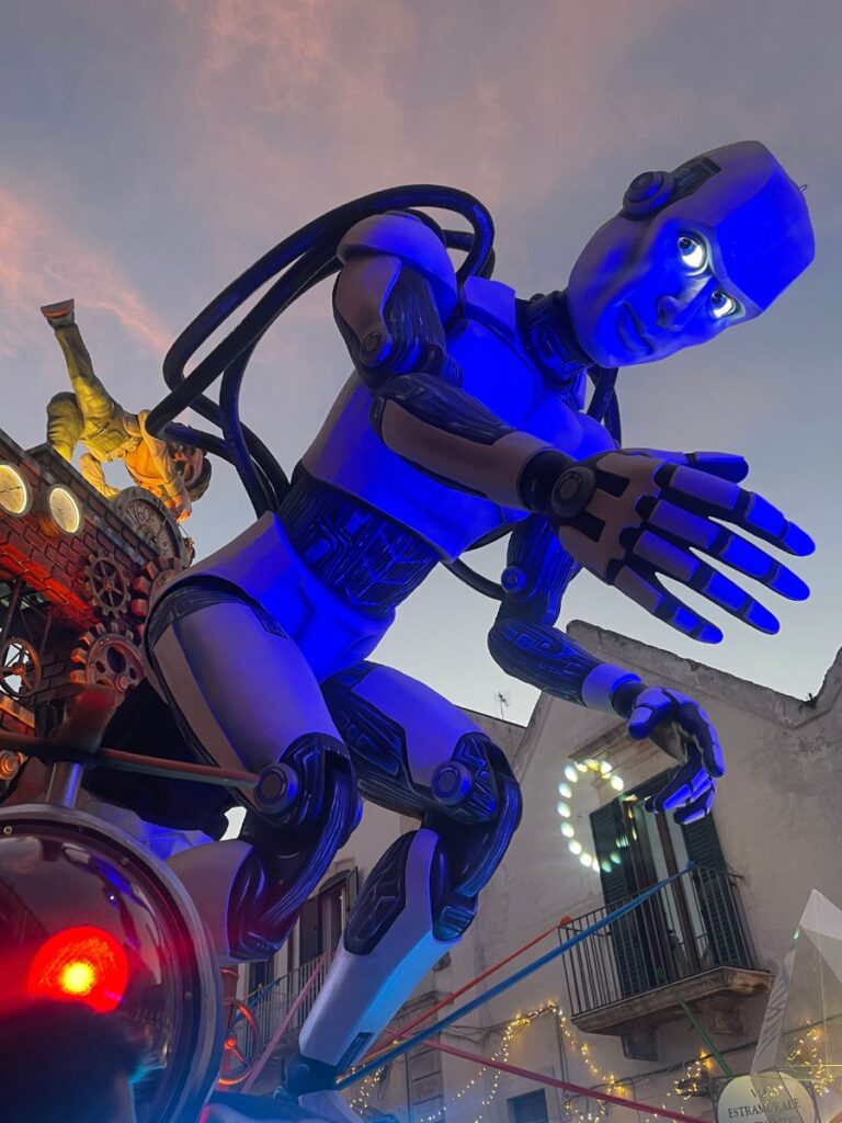 Una grande scultura di robot blu con occhi luminosi e arti meccanici si sporge in avanti contro un cielo serale scarsamente illuminato, creando una scena futuristica e accattivante.