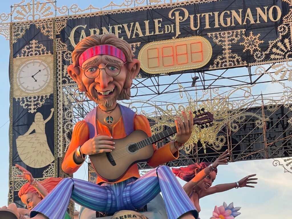 Un colorato carro da parata al Carnevale di Putignano presenta una grande e allegra figura hippie con occhiali e fascia, che suona la chitarra. Le decorazioni includono intricati disegni dorati e stendardi del festival.