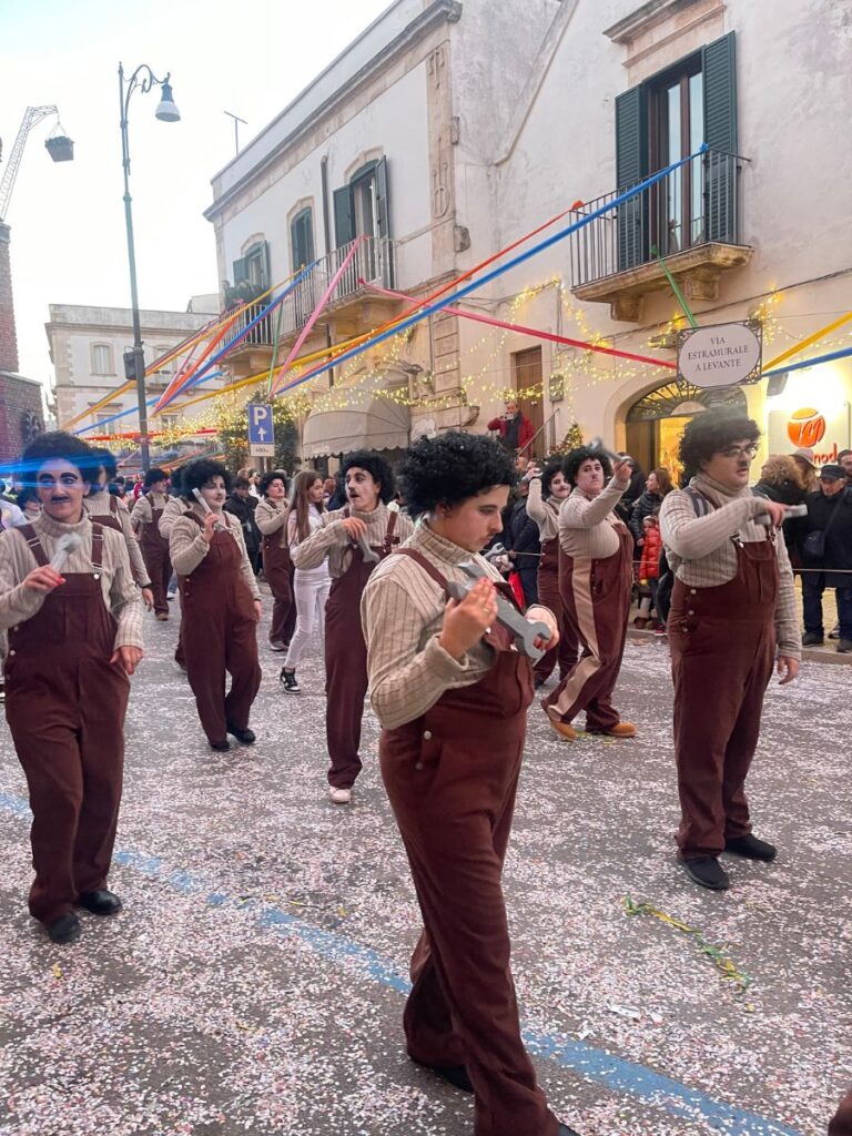 Una festosa parata di strada presenta persone in tuta marrone, parrucche nere e baffi, che camminano su una strada ricoperta di coriandoli. Nastri colorati pendono sopra.